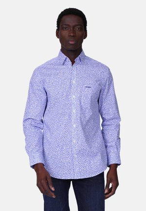 Homme portant une chemise boutonnée à manches longues blanche à motifs floraux bleus et un jean foncé, debout devant un fond uni.