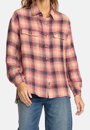 Kvinde iført en langærmet, ternet skjorte med knapper i pink, marineblå og creme med brystlommer foran, kombineret med blå jeans.