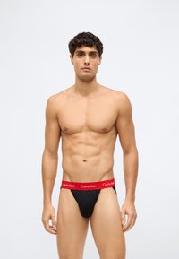Sorte truser med rød elastikk som har "Calvin Klein" logoen. Stretchy materiale, tettsittende design, minimal dekning med en jevn tekstur.