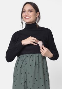 Czarna bluzka z długim rękawem i żebrowanym kołnierzykiem, nałożona na zieloną sukienkę w czarne kropki z guzikiem na przodzie.