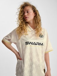 Vrouw met krullend haar die een crèmekleurig polo met patroon en het logo "SWARM International" draagt, kijkt zijwaarts met haar hand in de zij tegen een effen achtergrond.