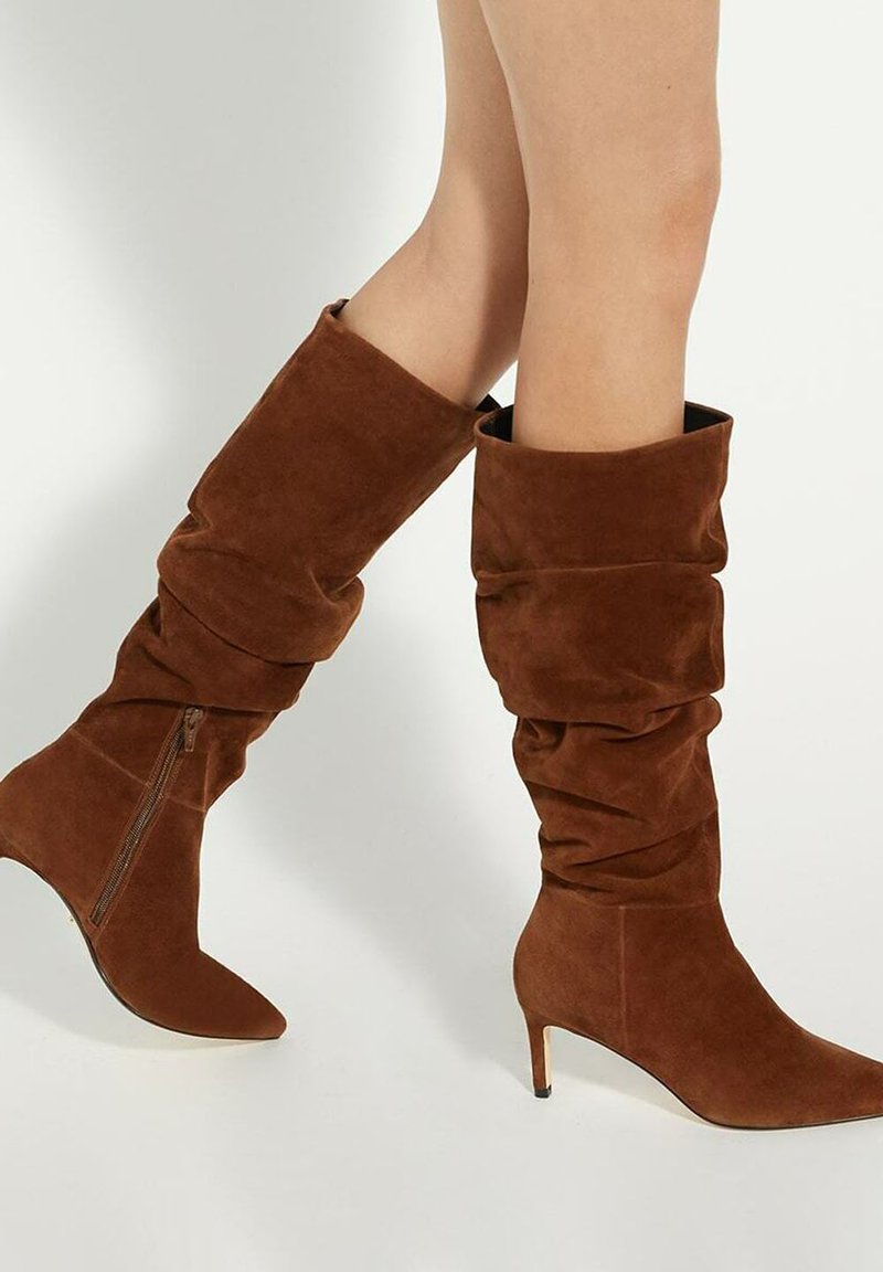 Bottes en daim marron jusqu'au genou avec un design décontracté, bout pointu et talon aiguille de 3 pouces. Dotées d'une fermeture éclair sur le côté pour un enfilage facile.