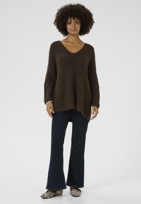 Brown knitted sweater with a V-neck, ribbed texture, and longer back hem, paired with dark blue flared jeans and black-and-white striped shoes.