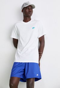Vit bomullströja med turkosa Nike-logotypen, kombinerad med blå shorts och en grå basebollkeps med broderade detaljer.