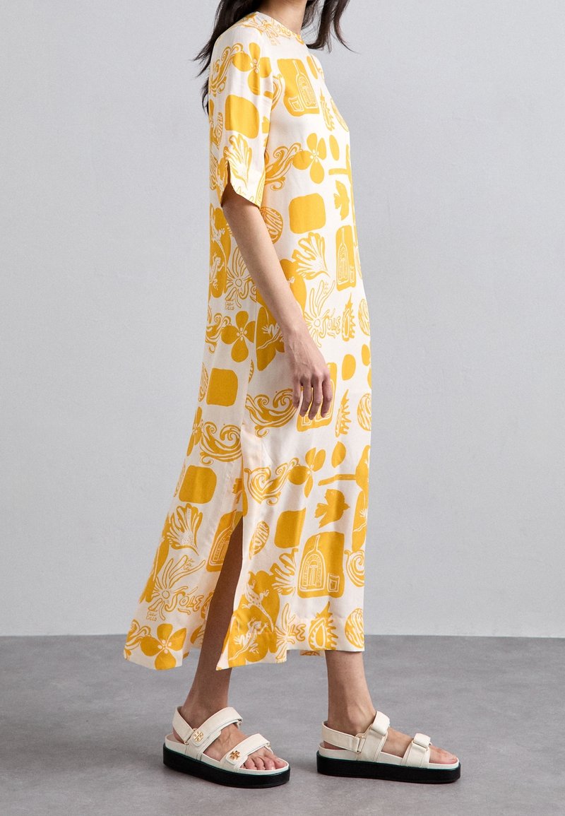 Robe midi jaune et blanche à motifs, avec manches courtes, fentes latérales, et sandales plates à semelle épaisse noire et brides crème.