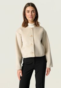 Beige cropped jacket in soft fabric with long sleeves and round neckline; features three buttons in front and a clean, minimalist design.