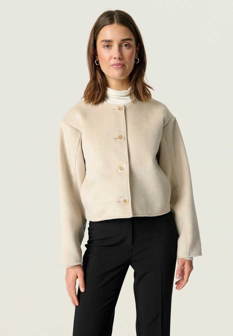 Beige cropped jacket in soft fabric with long sleeves and round neckline; features three buttons in front and a clean, minimalist design.