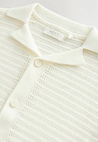 Pleteni beli srajc s polo ovratnikom, z teksturo in gumbi, ter oznako, ki prikazuje blagovno znamko in izvor.