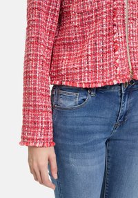 Czerwony żakiet z wełny tweedowej o teksturowanym splocie, z frędzlami na mankietach i zamkiem błyskawicznym, w połączeniu z niebieskimi dżinsami z przednią kieszenią.