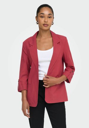 Femme aux cheveux foncés en chignon bas, vêtue d'un blazer rouge sur un haut blanc et un pantalon noir, debout devant un fond clair uni.