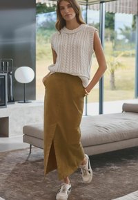 Pletený bílý top bez rukávů s texturovaným vzorem, spárovaný s dlouhou olivově zelenou sukní s předním rozparkem, a bílými teniskami.
