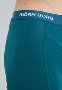 Tirkizne pamučne bokserice s plavim elastičnim pojasom na kojem piše "BJÖRN BORG" bijelim slovima. Glatka tekstura i prilagođen dizajn s bešavnim stranama.