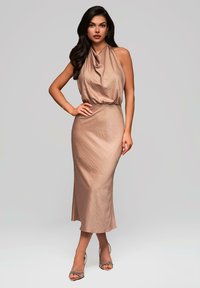 Robe à col halter en tissu beige clair, avec un corsage drapé et une jupe ajustée arrivant au genou. Des talons à lanières complètent le look.