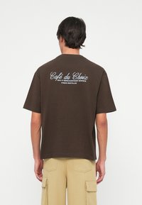 Καφετί βαμβακερό T-shirt με κοντά μανίκια. Η πλάτη διαθέτει λευκό εκτυπωμένο κείμενο: "Café du Choix 24/7 Breakfast Diner Free Refills."