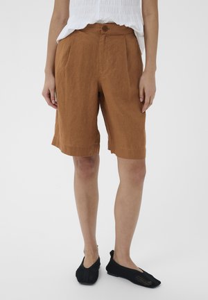 Žena má na sebe hnedé šortky s vysokým pásom po kolená s predným gombíkom a čierne sieťované slip-on topánky s rozparkom, stojí pred jednoduchým pozadím.