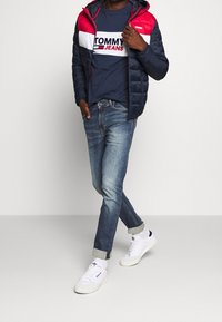 Fjäderjacka med röda, vita och marinblå paneler, bärs över en marinblå Tommy Jeans logot-skjorta, i kombination med ljusblå jeans och vita sneakers.