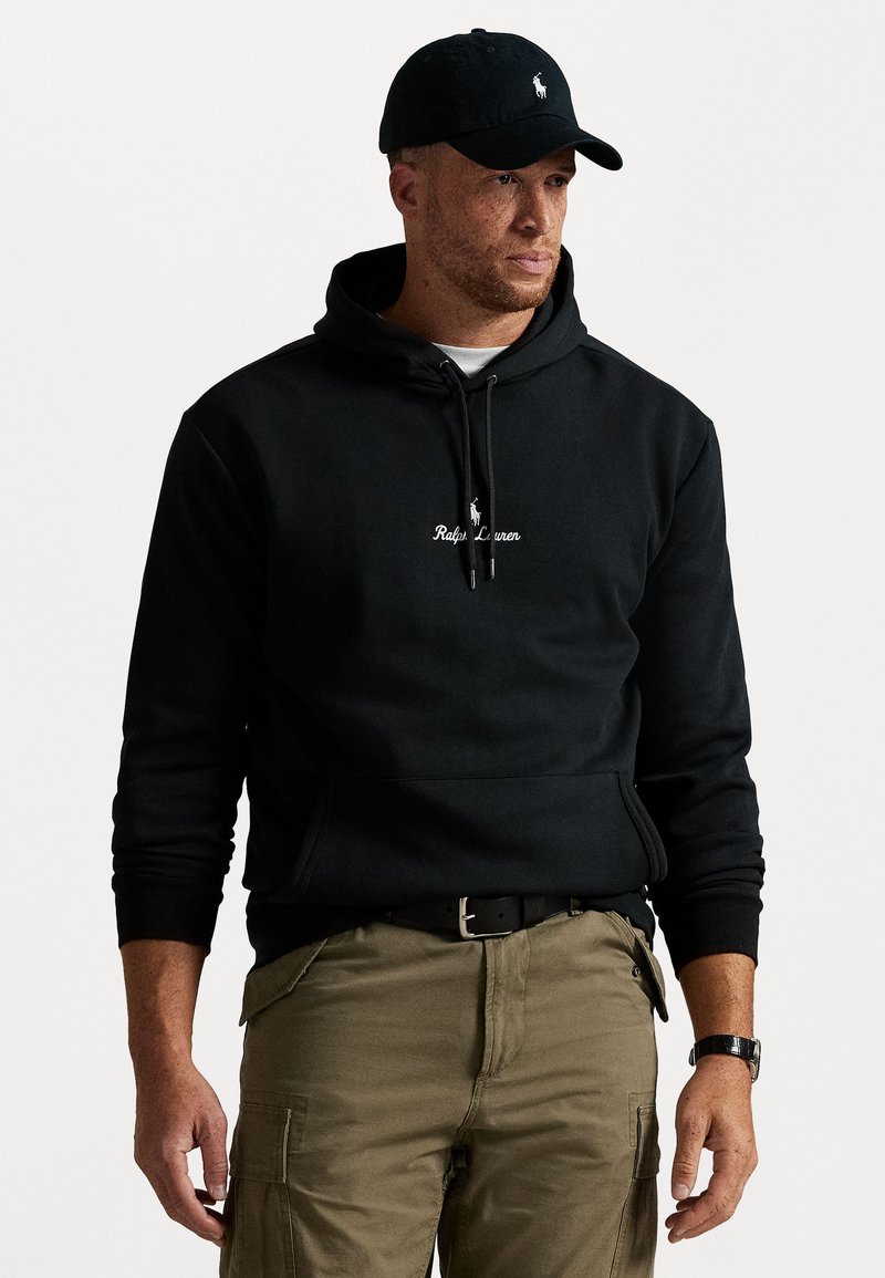 Mężczyzna ubrany w czarną bluzę z kapturem Ralph Lauren i czapkę, oliwkowe bojówki oraz zegarek na nadgarstku, stojący lekko zwrócony w prawo na białym tle.