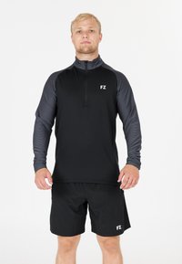 En blond man står vänd framåt iklädd en svart och grå långärmad zip-up topp och svarta shorts med vita "FZ"-logotyper.