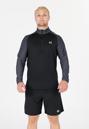 En blond mand står Vendt fremad iført en sort og grå langærmet zip-up trøje og sorte shorts med hvide "FZ" logoer.