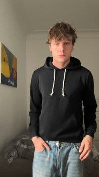 Un jeune homme avec des cheveux en désordre portant un sweat à capuche noir et un jean bleu clair se tient dans une chambre avec des œuvres d'art abstrait et un lit en arrière-plan.