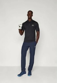 Μαύρο πόλο μπλουζάκι με λογότυπο, ναυτικές παντελόνες και μπλε αθλητικά παπούτσια. Ο άνδρας κρατά ένα μπαστούνι γκολφ φορώντας λευκά γάντια γκολφ. Απλός, μοντέρνος σχεδιασμός.