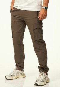 Braune Cargo-Hosen aus einem glatten Stoff, mit Seitentaschen, elastischen Bündchen und schlankem Schnitt. Tragen Sie sie mit grauen Turnschuhen.