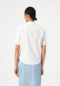 Femme aux cheveux tressés portant une chemise blanche à manches courtes et une jupe en jean bleu clair, vue de dos sur un fond blanc uni.
