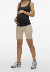 Top de maternidad sin mangas en negro, combinado con pantalones cortos de ciclismo beige, con un diseño ajustado. La modelo lleva zapatillas deportivas plateadas.
