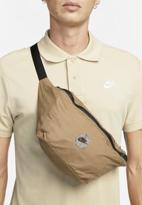 Hnědá polstrovaná taška přes rameno s černým zipem, zdobená kulatou nášivkou s logem Nike. Nastavitelný popruh a hladká textura.