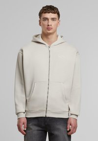 Lys beige zip-up hette i myk bomullsblanding, med kengurulomme, ribbestrikkede mansjetter og en liten brodert logo på brystet.