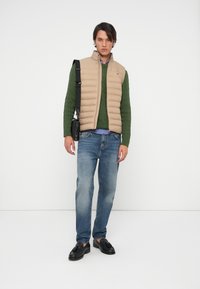Beige puffer vest over green cable-knit sweater, blue checkered shirt, blue jeans, and black loafers. Minimalist crossbody bag on shoulder.