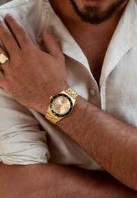 Goldene Uhr mit einem strukturierten Metallband, rundem Ziffernblatt und schwarzer Lünette. Das Zifferblatt hat silberne Akzente und Zahlenmarkierungen. Getragen an einem gebräunten Handgelenk.