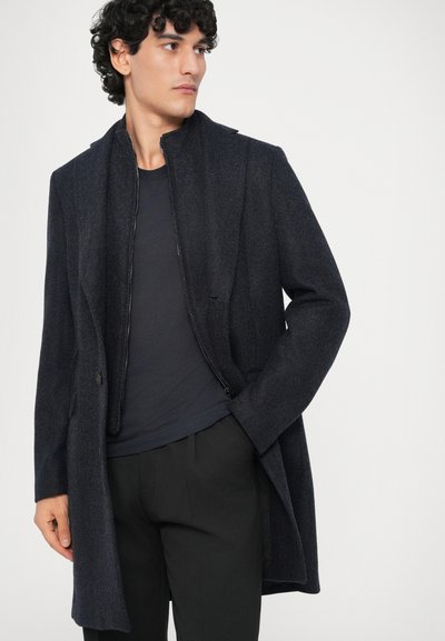 Hombre vestido con un abrigo de lana azul marino oscuro sobre una chaqueta con cremallera y una camisa azul marino, con pantalones negros, mirando hacia un lado con una mano en el bolsillo.