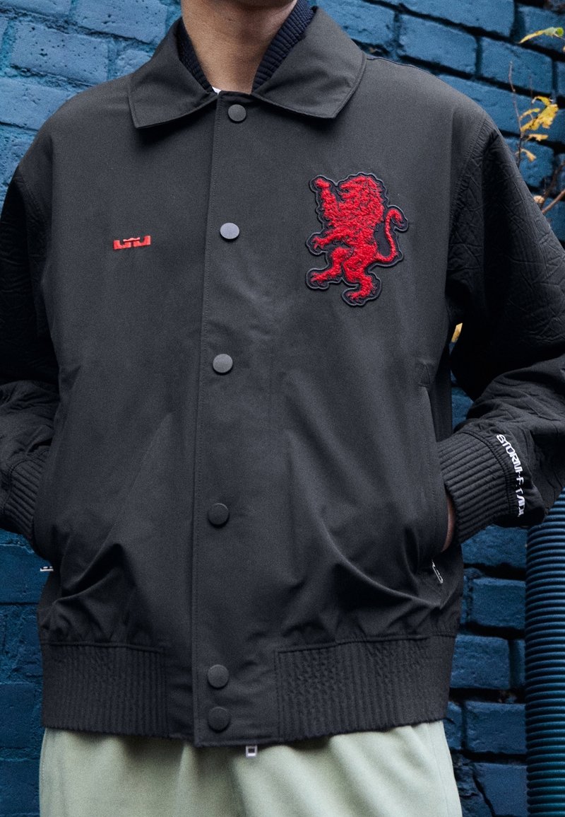 Veste bomber noire avec un emblème de lion rouge brodé, boutons-pression, poignets côtelés et texture matelassée sur les manches.