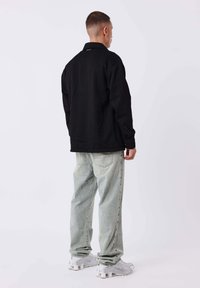 Chaqueta negra texturizada con cuello tipo solapa, combinada con jeans de corte holgado de lavado claro y zapatillas blancas, vista desde la parte trasera.