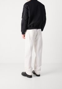 Veste bomber noire matelassée, pantalon blanc ample et bottes noires. La veste possède un col côtelé ; le pantalon a des poches arrière.