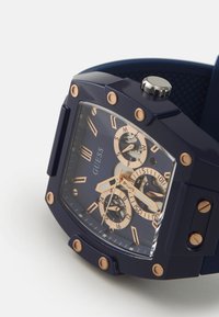 Montre pour homme avec un bracelet en silicone bleu marine, un cadran carré, des accents en or rose, des sous-cadrans chronographes et un affichage transparent.