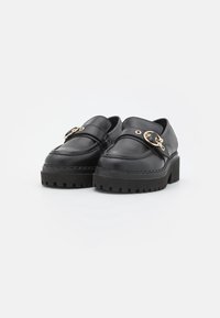 Zapatos de plataforma de cuero negro con una suela gruesa, que presentan un hebilla dorada decorativa y un detalle de costura elegante.