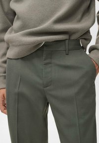 Suéter de punto gris superpuesto sobre pantalones oscuros verdes, ajustados, con bolsillos laterales, textura suave y diseño de frente plano.