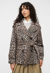 Μπεζ και μαύρο παλτό με τύπωμα λεοπάρδαλης, με διπλό πέτο, φαρδιά γιακά και ζώνη από ύφασμα. Φορεμένο πάνω από ένα ελαφρύ πουλόβερ.