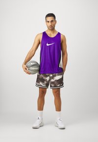 Lillasest Nike'i tops, mustade ja valgete mustritega lühikesed püksid, valged tossud, hoiab korvpalli. Topsil ja pükstel on logo.