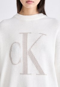 Bela pletena jopica z velikim, svetlo rjavim logotipom "CK" na sredini. Tkanina ima teksturirano, odprto tkanje. Vidna rebrasta ovratnica.