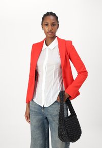 Blazer rouge ajusté sur une chemise blanche à boutons, associé à un jean en denim à jambes larges. Accessoirisé avec un sac à main noir texturé.