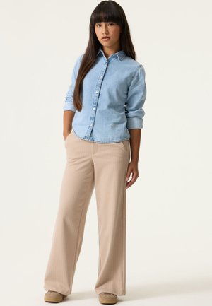 Frau mit langen dunklen Haaren, die ein hellblaues Jeanshemd, beige weit geschnittene Hosen und beige Schuhe trägt, steht mit den Händen in den Taschen.