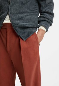 Grå ribbad stickad cardigan med dragkedja, ihop med rostfärgade skräddarsydda byxor med slät yta och framfickor.