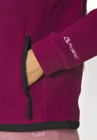 Fleecejacka i djup magenta med svarta detaljer; har en ficka med dragkedja och Polartec-logotyp på ärmen. Mjuk textur och figurnära design.