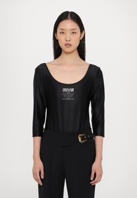 Bodysuit negro con mangas de tres cuartos, hecho de un tejido brillante. Presenta un escote redondeado y un logo impreso en la parte frontal. Combinado con pantalones de talle alto.