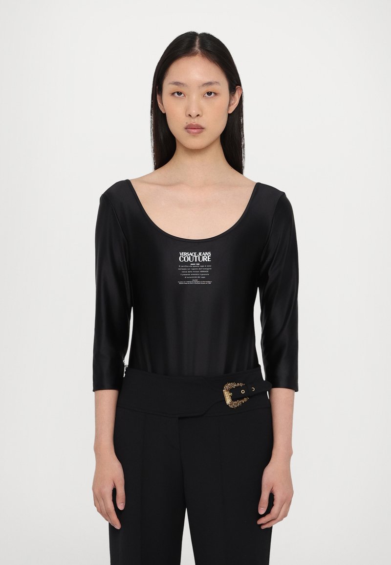 Bodysuit negro con mangas de tres cuartos, hecho de un tejido brillante. Presenta un escote redondeado y un logo impreso en la parte frontal. Combinado con pantalones de talle alto.