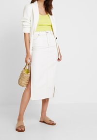 Femme portant un blazer blanc, un haut sans manches jaune, une jupe midi blanche avec une fente, des sandales glissantes beige, et tenant un sac en osier.