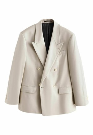 Person adjusting the sleeve of a cream blazer worn over a blue denim shirt against a plain background.